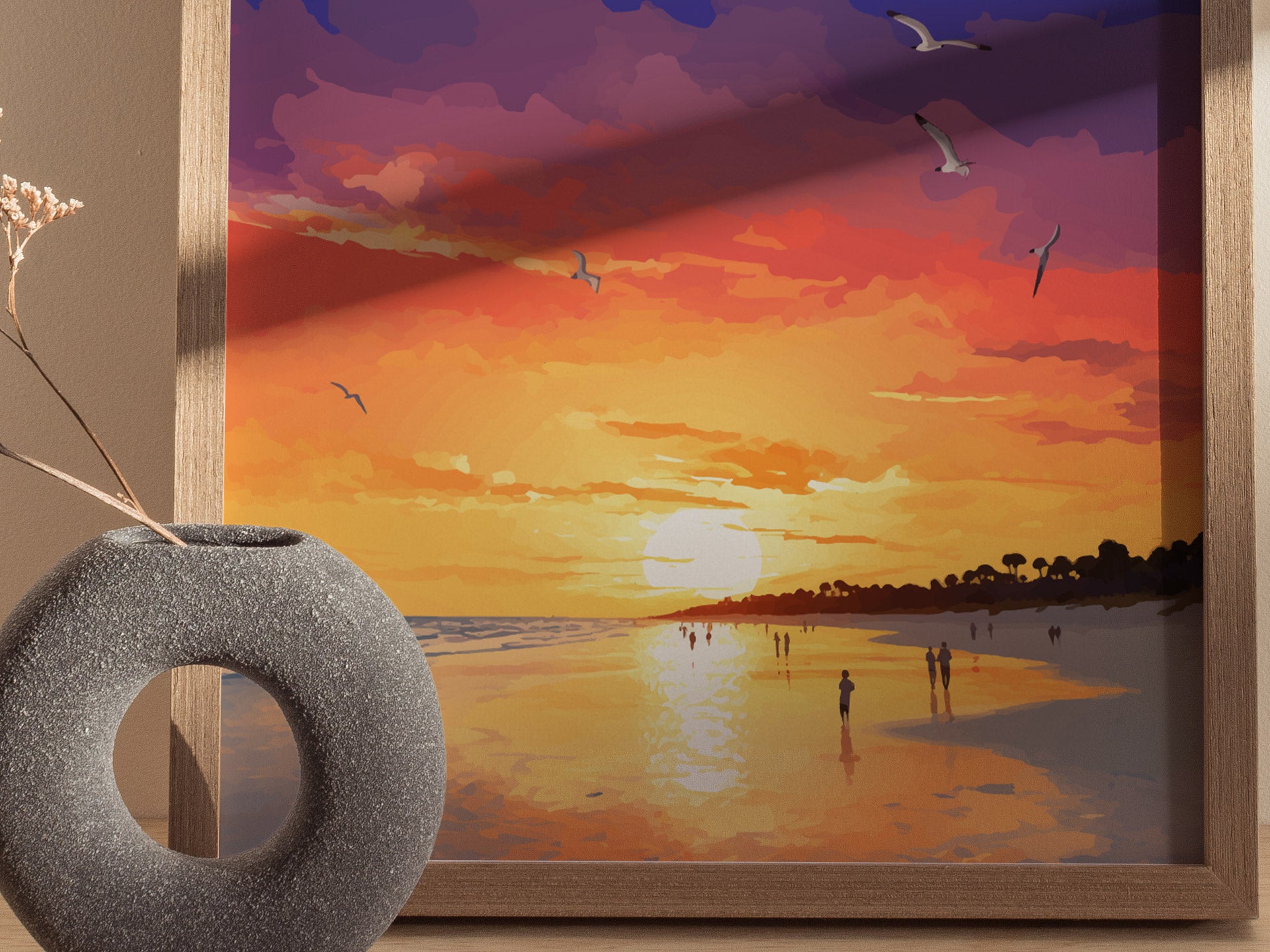 A framed poster depicts a vibrant sunset with purples, oranges, and yellows over a beach, featuring silhouettes of people and seagulls. A round, textured vase sits nearby.