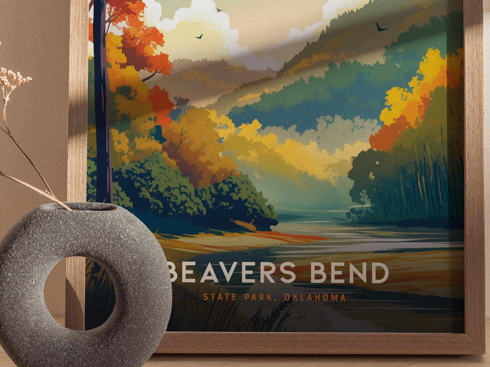 Framed wall art of Beavers Bend State Park showcases colorful autumn foliage and a serene river landscape. A round, textured gray vase with dried flowers sits in the foreground.