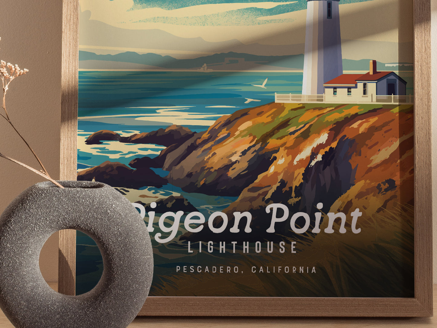 A framed poster of Pigeon Point Lighthouse shows a vibrant coastal scene with cliffs and ocean waves. A gray circular vase with dried flowers is in the foreground.