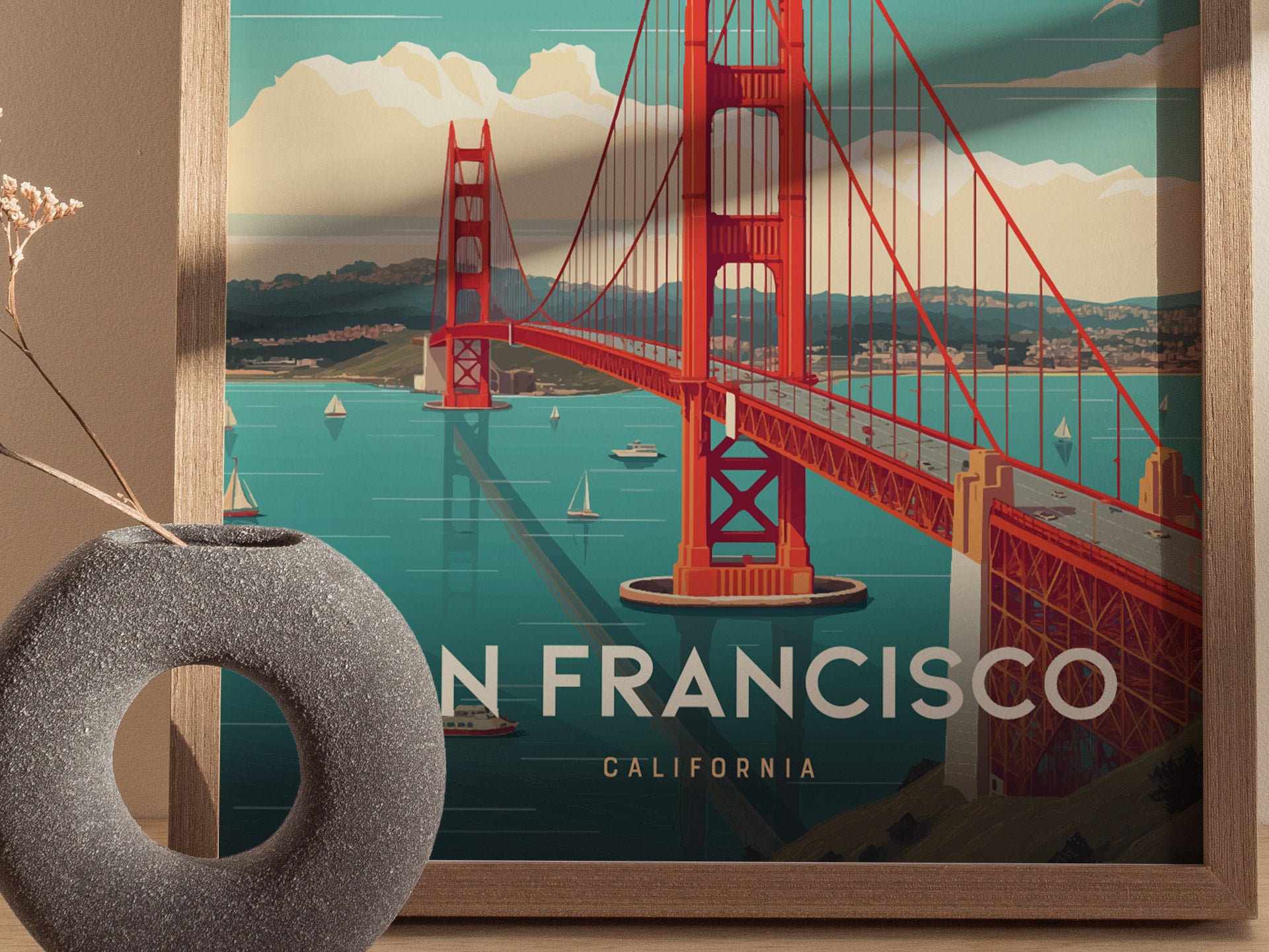 A vibrant poster of the Golden Gate Bridge with sailboats in the bay, framed in wood. A decorative stone vase with dried flowers sits in the foreground.