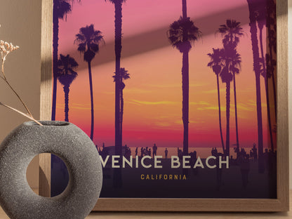 A framed Venice Beach poster featuring silhouettes of palm trees against a vibrant pink and orange sunset. A round stone vase with dried flowers is placed in the foreground.