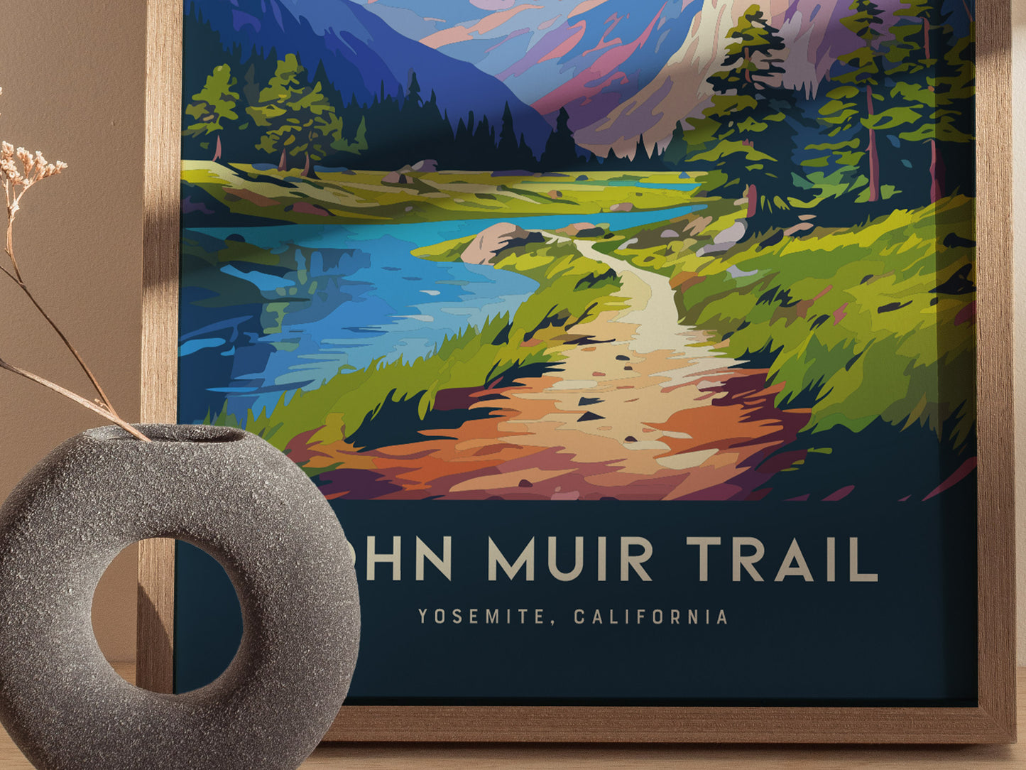 A colorful John Muir Trail poster depicts a scenic path with lush greenery, a tranquil blue lake, and distant mountains. A textured round vase with dried flowers accents the composition.