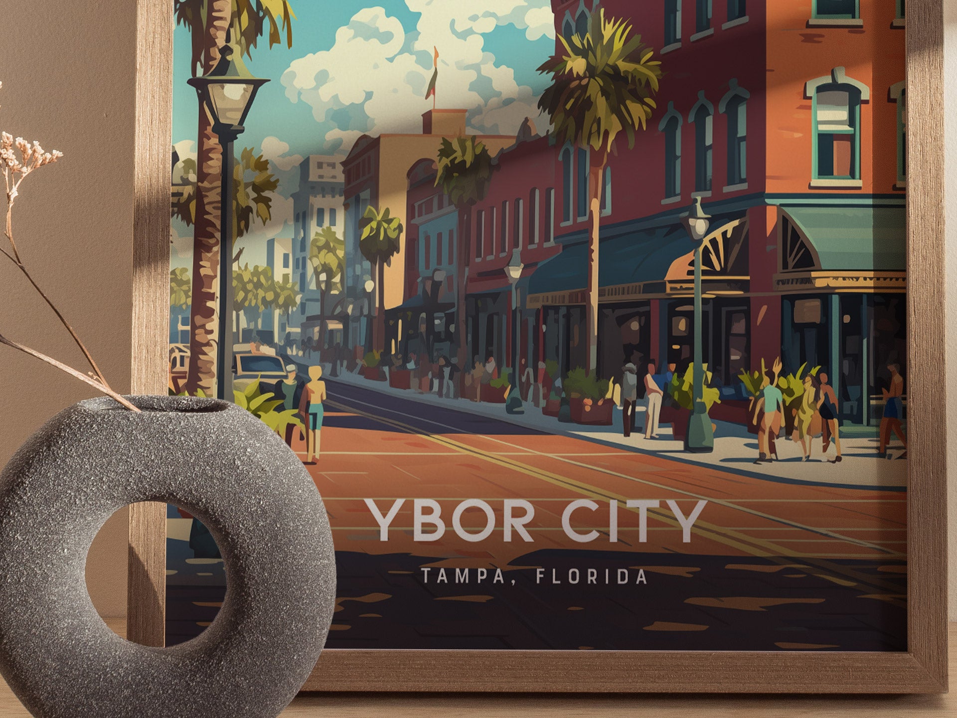 A colorful poster of Ybor City, Tampa, Florida, featuring vibrant street scenes, palm trees, and historic architecture. A grey circular vase with dried branches is in the foreground.