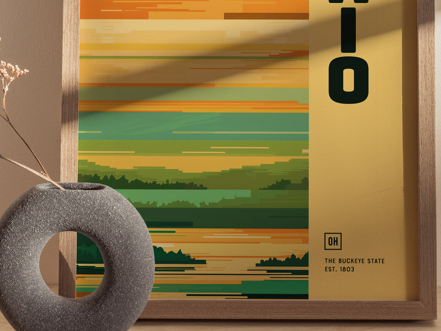 A framed Ohio poster features horizontal green and yellow lines with The Buckeye State Est. 1803 text. A round gray vase with dried flowers is placed in the foreground.
