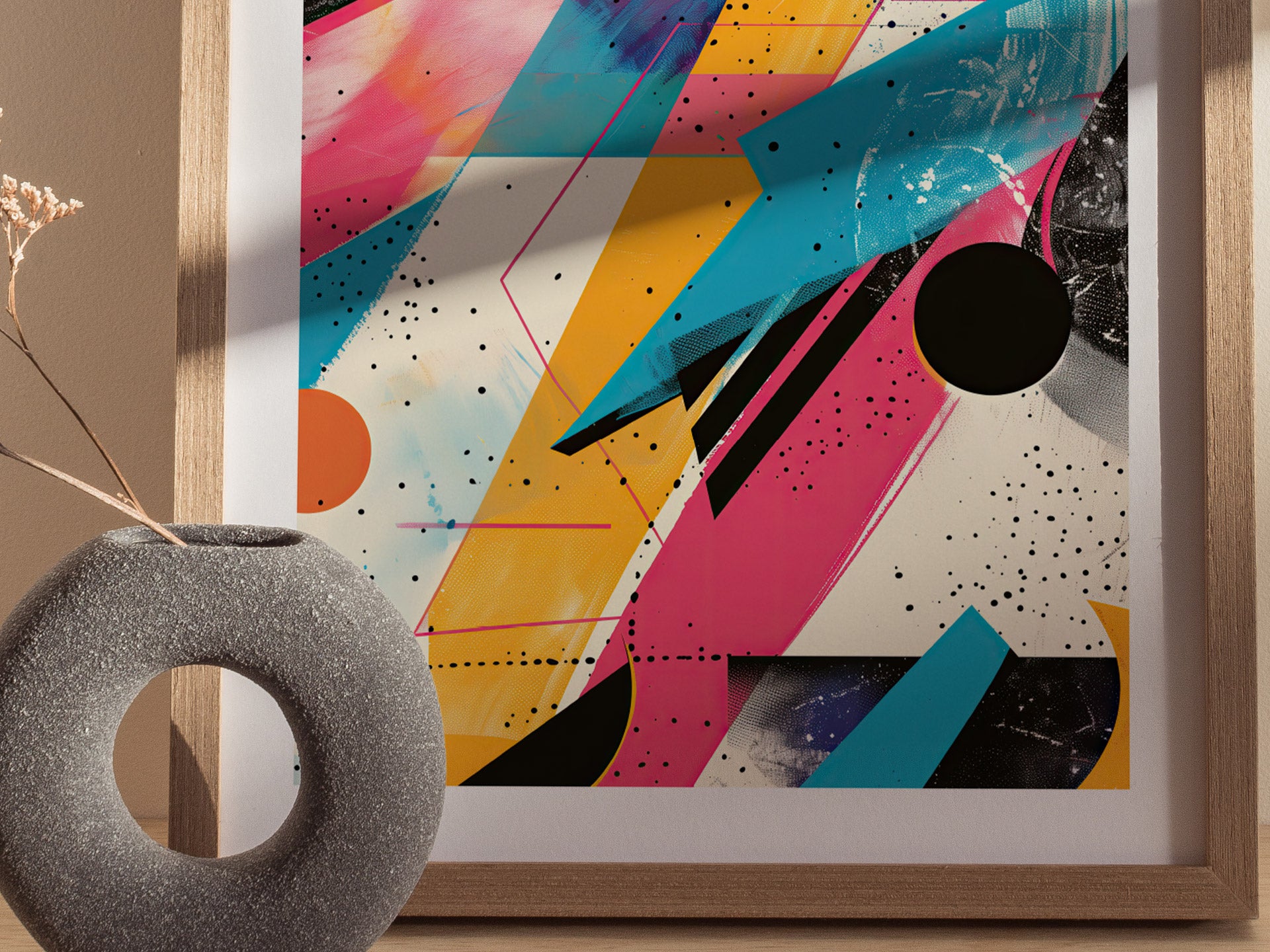 A framed 1980s-style abstract poster features vibrant geometric shapes in pink, blue, and yellow with black speckles. A textured gray vase with dried flowers is visible in the foreground.