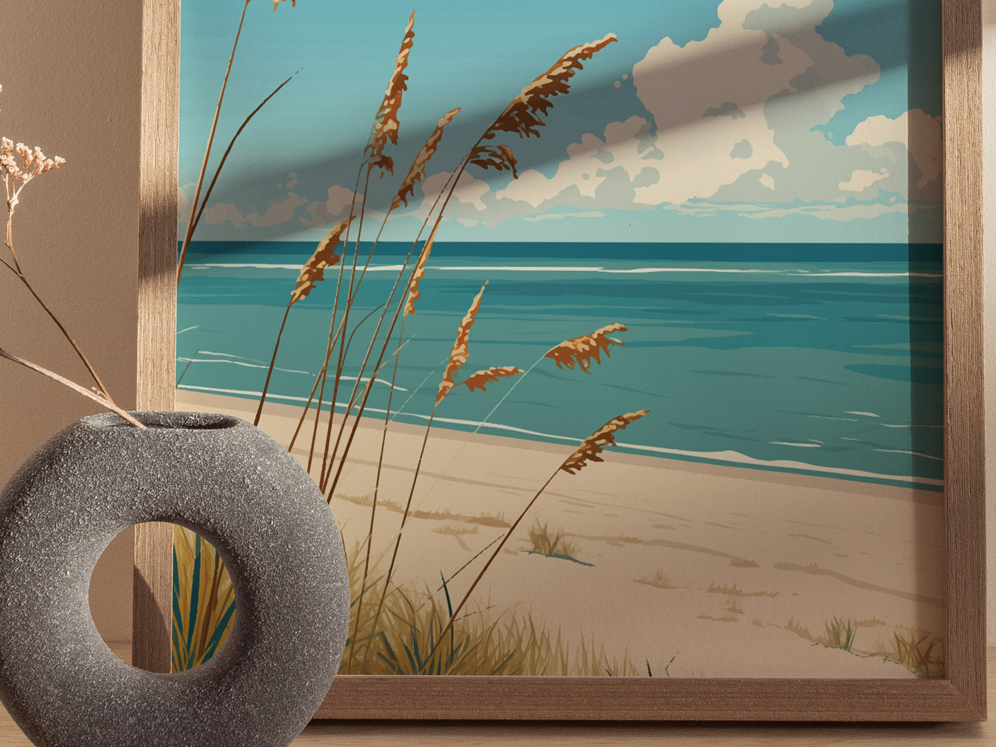 A framed poster depicting a serene beach scene with turquoise waves and tall grasses is partially visible behind a textured, circular gray vase holding dried flowers.