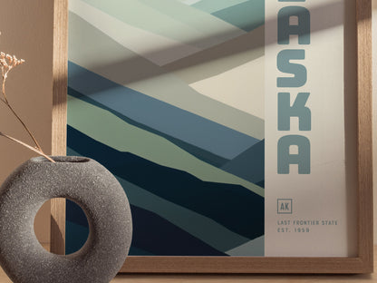 A framed minimalist poster featuring abstract mountain shapes in shades of blue and green, with ALASKA and Last Frontier State Est. 1959 text. A round, textured vase sits nearby.
