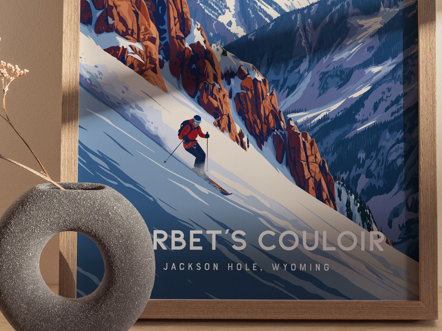 A framed poster of a skier descending Corbets Couloir, surrounded by rugged snow-covered mountains, is displayed next to a round, textured vase with dried twigs.