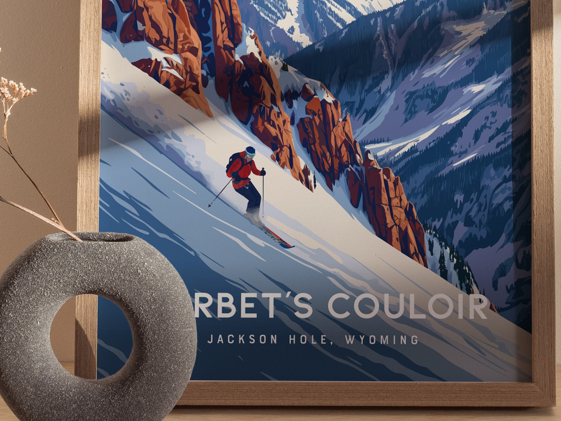 A framed poster of a skier descending Corbets Couloir, surrounded by rugged snow-covered mountains, is displayed next to a round, textured vase with dried twigs.