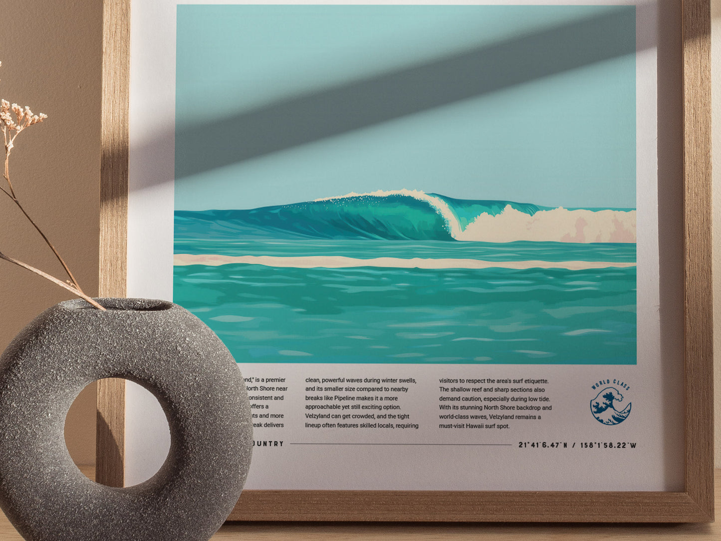 Framed poster depicting a turquoise ocean wave, with text and coordinates below, set against a beige wall. A round, textured vase is in the foreground.