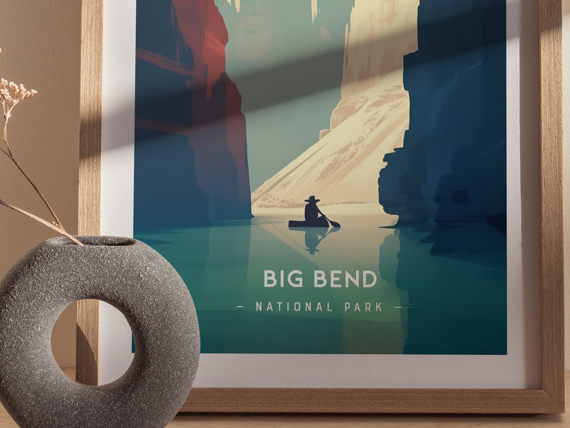 A framed minimalist poster of Big Bend National Park depicts a lone canoeist on a calm river between towering cliffs. A textured gray vase with dried flowers is nearby.