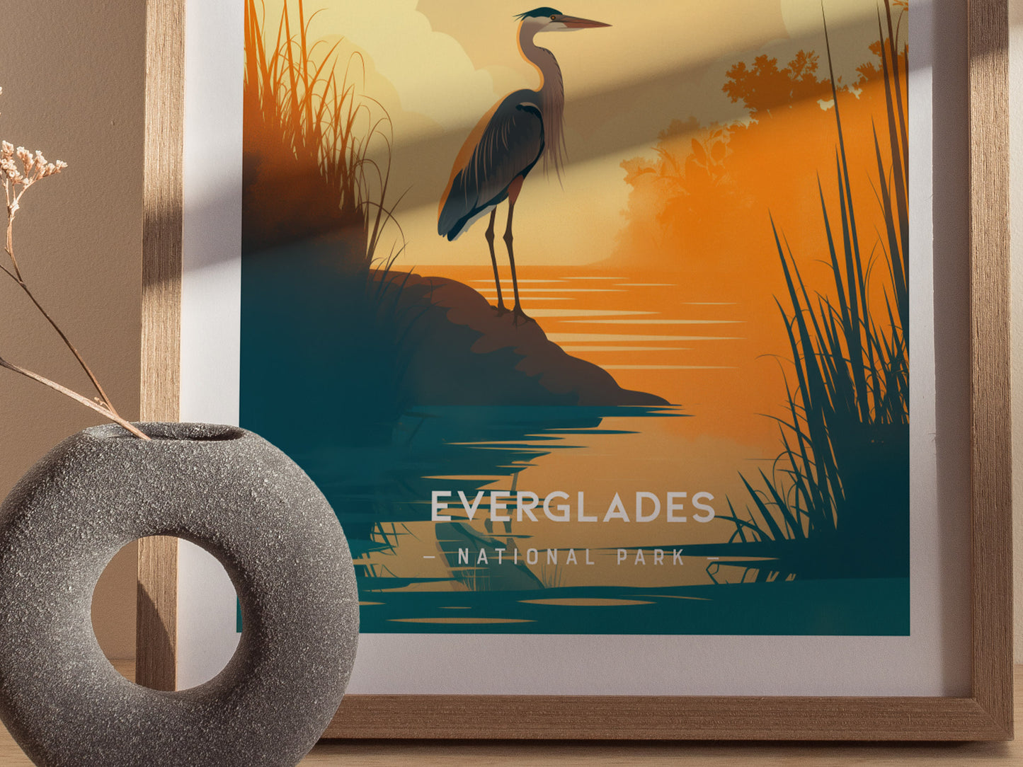 A framed minimalist poster of the Everglades features a heron by the water at sunset, with tall grasses and warm orange and blue hues. A round vase is nearby.