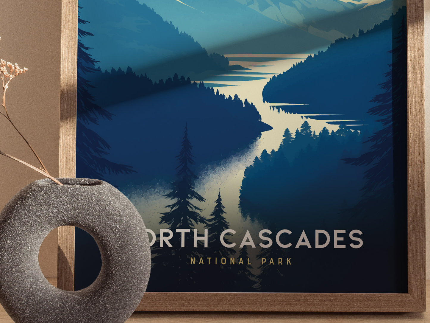 A framed poster of North Cascades National Park featuring stylized mountains, trees, and a winding river. A textured round vase with dried flowers is placed in the foreground.