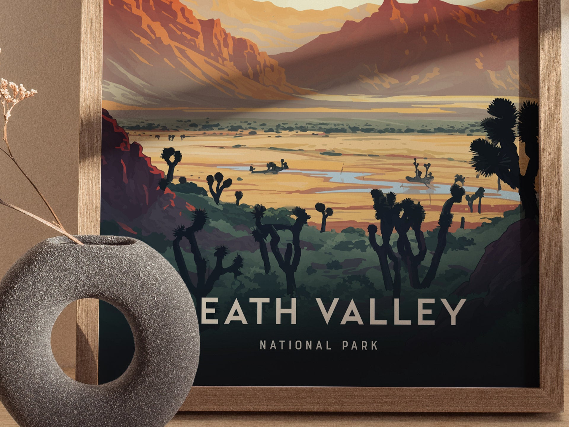 A framed print depicting a stylized landscape of Death Valley with desert plants and distant mountains, placed beside a textured round vase with dried flowers.
