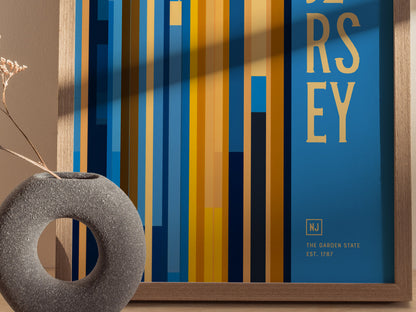A New Jersey-themed poster with vertical blue and yellow stripes and text, framed in wood, is displayed next to a textured circular vase with dried flowers.