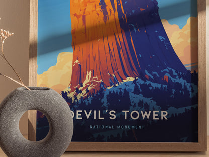 A colorful poster of Devils Tower depicts the geological formation in vibrant oranges and blues. The artwork is framed alongside a textured, circular gray vase on a wooden surface.
