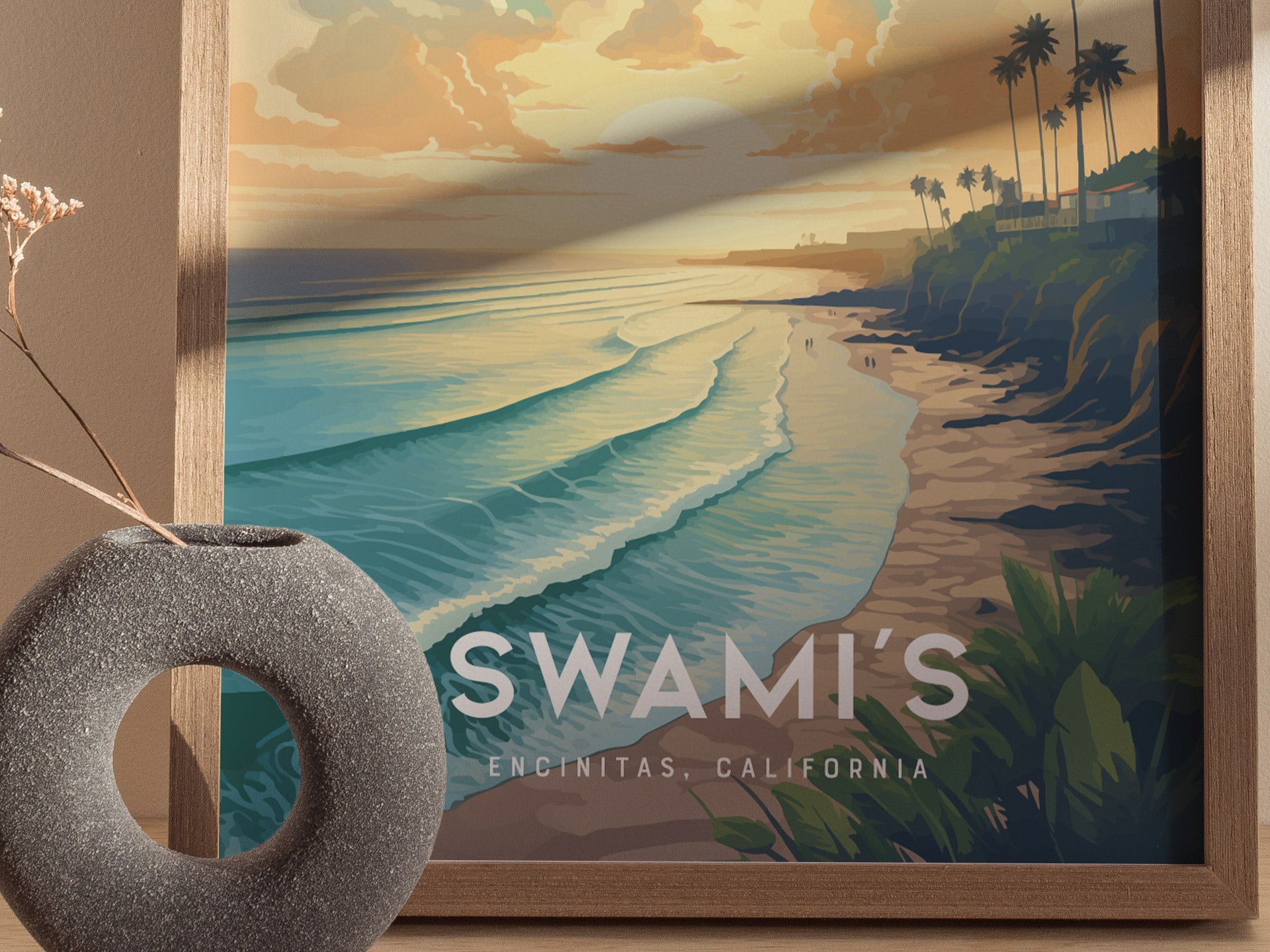 A framed poster depicts a California beach at sunset with waves rolling in and palm trees silhouetted on a cliff. A textured gray vase sits nearby.