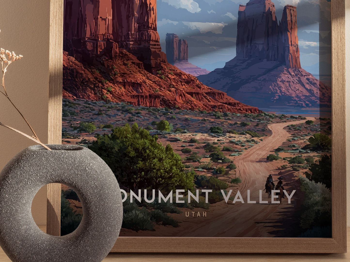 A framed poster of Monument Valley features red sandstone mesas under a cloudy sky, with a winding dirt road and two riders on horseback.