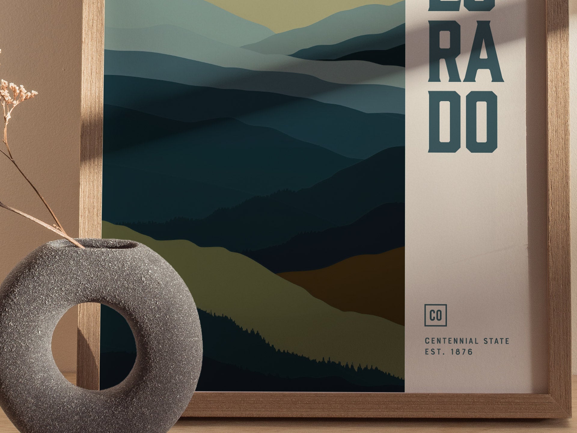 A framed minimalist poster depicting abstract layers of blue and green mountains with Colorado text. A textured, circular vase with dried flowers is placed in the foreground.
