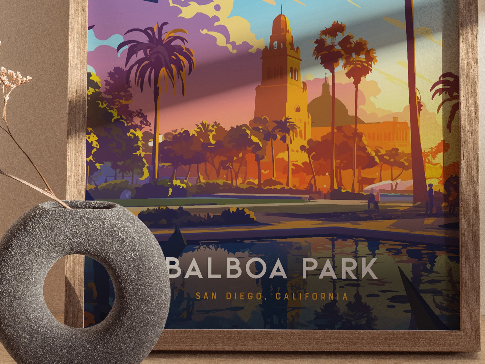 A framed art print depicts a vibrant, colorful illustration of Balboa Park with palm trees and Spanish Colonial architecture. A circular stone vase with dried flowers is in the foreground.