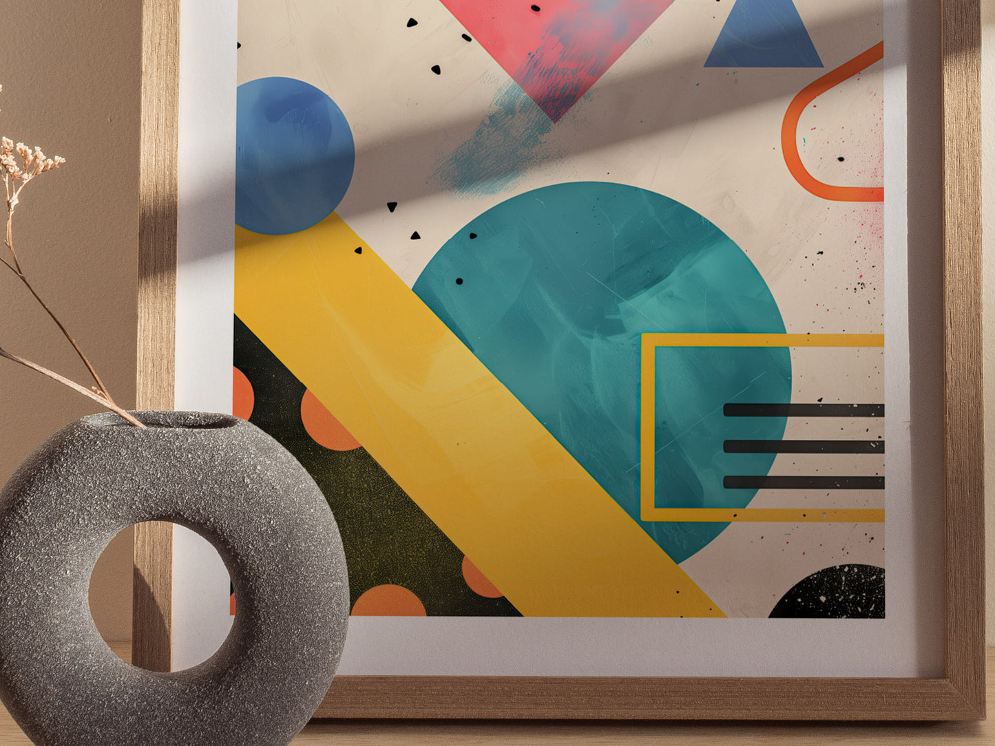 A framed abstract art piece features bold geometric shapes and vibrant colors, including teal circles and yellow triangles. A textured gray circular vase sits in the foreground.