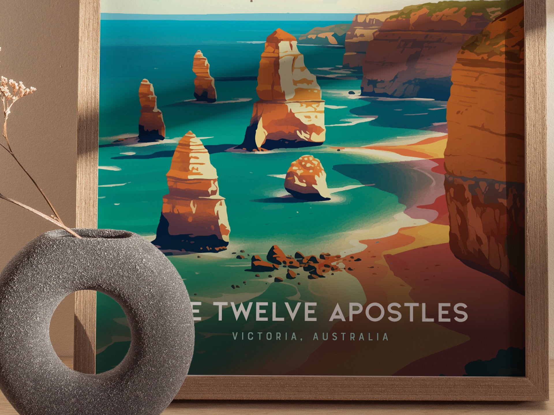 A framed poster depicts the Twelve Apostles, showcasing vibrant turquoise waters and towering limestone stacks along the coast. A modern, round vase with dried flowers sits nearby.