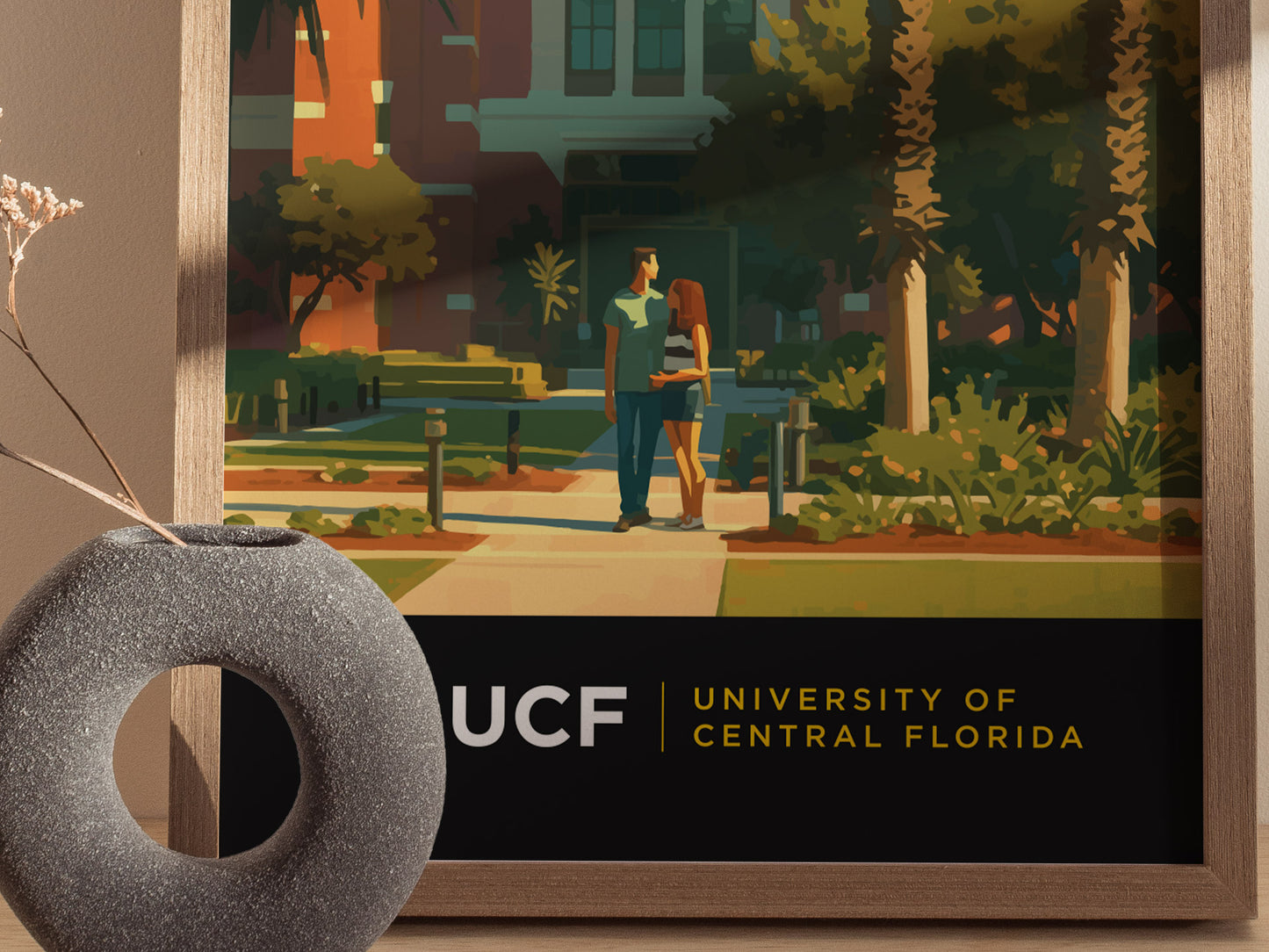 A framed poster of the University of Central Florida campus features two people walking among palm trees. The foreground includes a modern vase with dried flowers.
