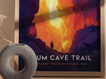 A colorful poster depicts a hiker on the Alum Cave Trail surrounded by vibrant orange and purple rock formations. A textured gray vase with dried flowers is in the foreground.