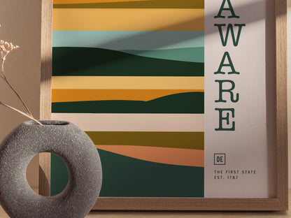 A minimalist Delaware-themed poster with layered abstract shapes in warm earthy tones is framed on a wall. A round stone vase with dried flowers is placed in the foreground.