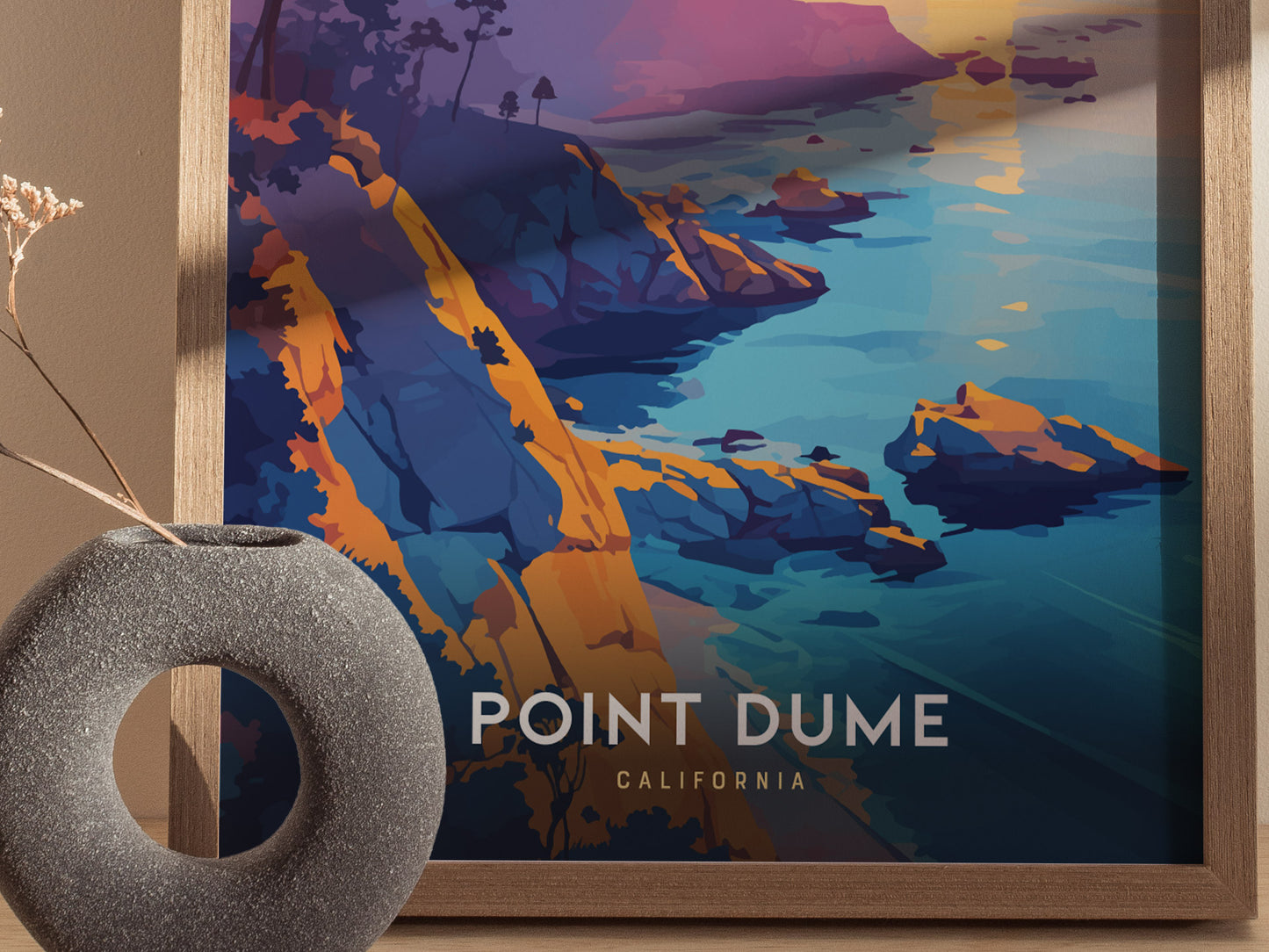 A framed poster of Point Dume, California, showcasing colorful cliffs and ocean in a modern art style, sits beside a round, textured vase with a dried plant.