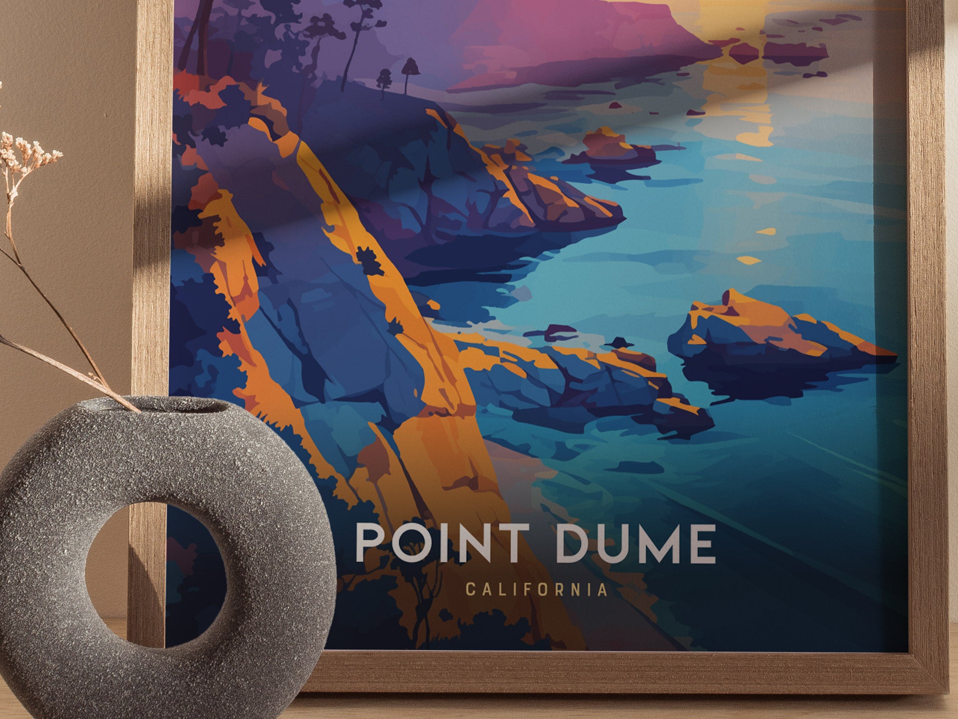 A framed poster of Point Dume, California, showcasing colorful cliffs and ocean in a modern art style, sits beside a round, textured vase with a dried plant.
