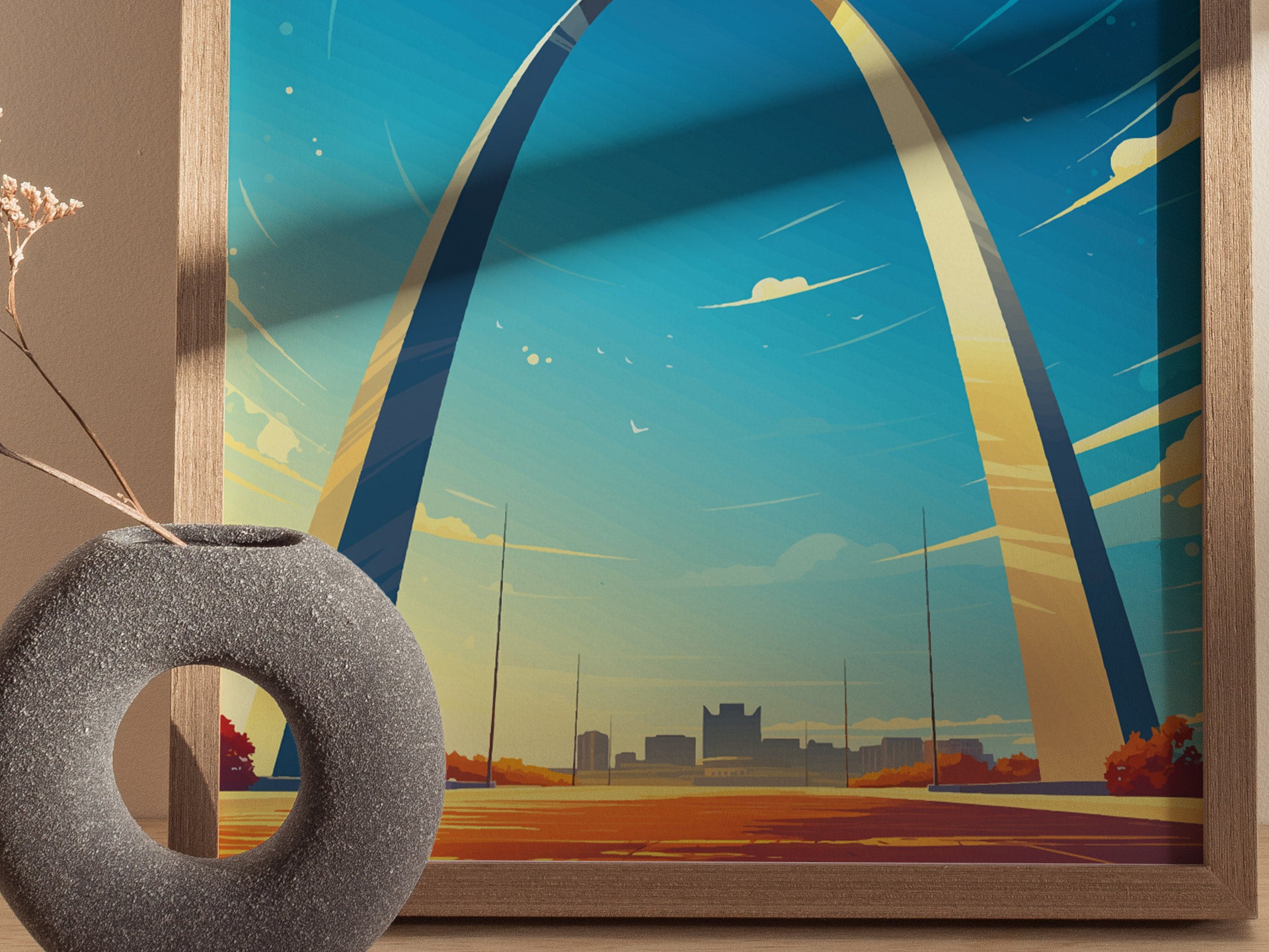 A framed poster of the Gateway Arch with a minimalist design, set against a vibrant blue sky, alongside a textured, circular vase with dried flowers.