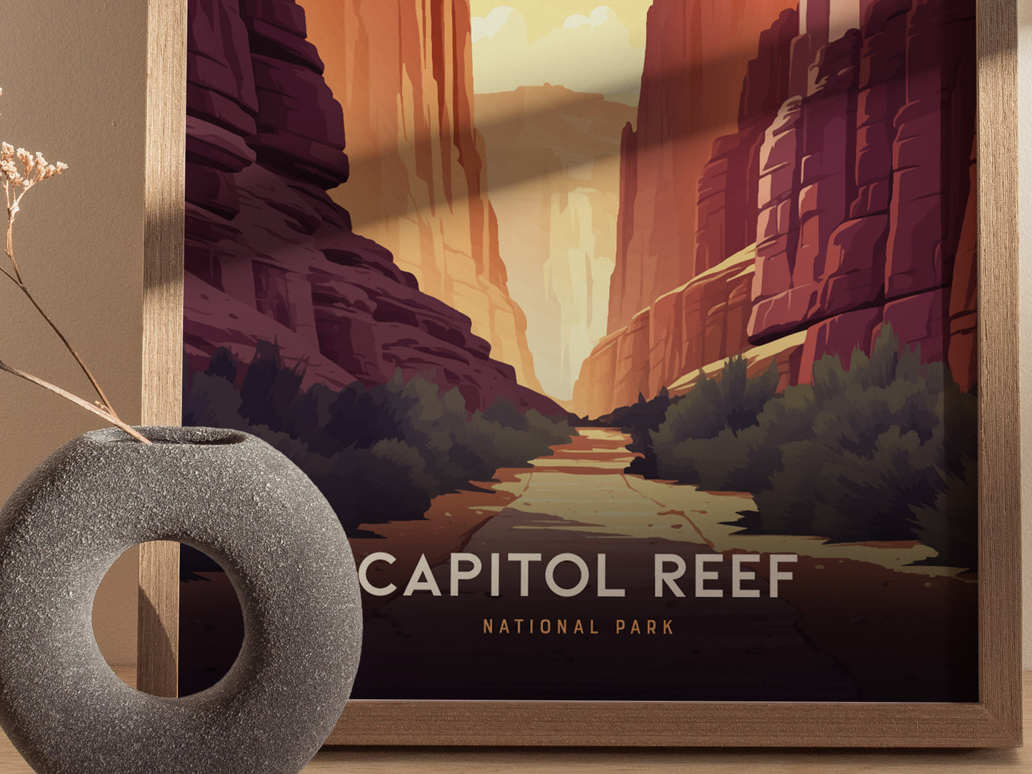 A framed art print depicting Capitol Reef National Park features towering red rock formations and a winding canyon path, with a textured grey vase in the foreground.
