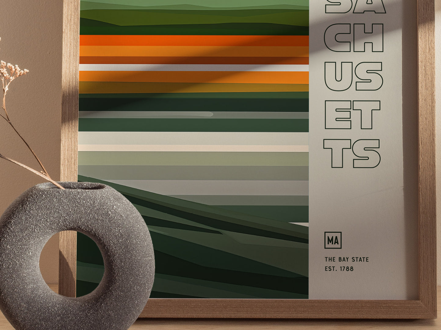 A framed Massachusetts poster features horizontal green and orange stripes with SACHUSETTS text. A round, textured vase with dried flowers is placed beside it.
