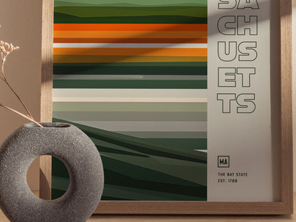 A framed Massachusetts poster features horizontal green and orange stripes with SACHUSETTS text. A round, textured vase with dried flowers is placed beside it.