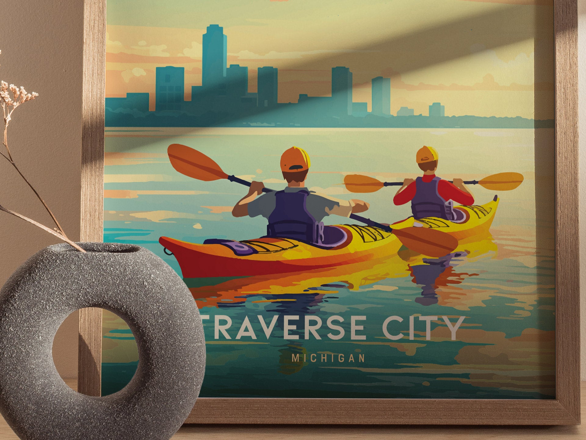 This vibrant poster features two kayakers paddling on calm waters with a city skyline in the background, framed in wood. A textured vase with dried flowers is nearby.