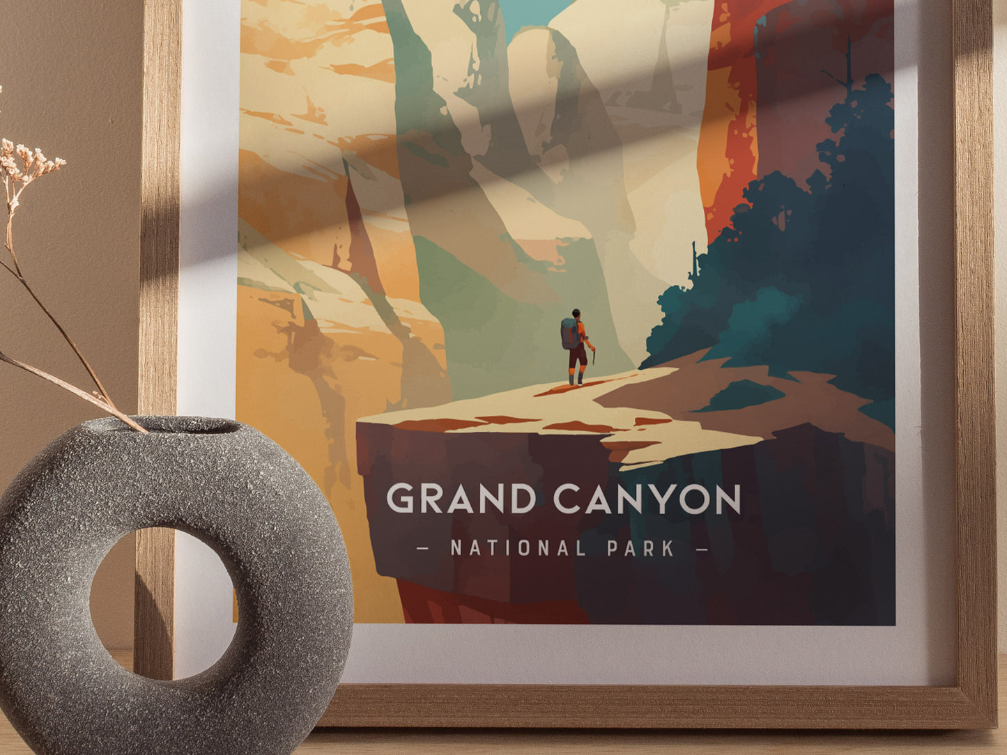 A framed minimalist illustration depicts a lone hiker overlooking the Grand Canyon with striking orange and teal tones. A textured round vase with dried flowers is placed nearby.