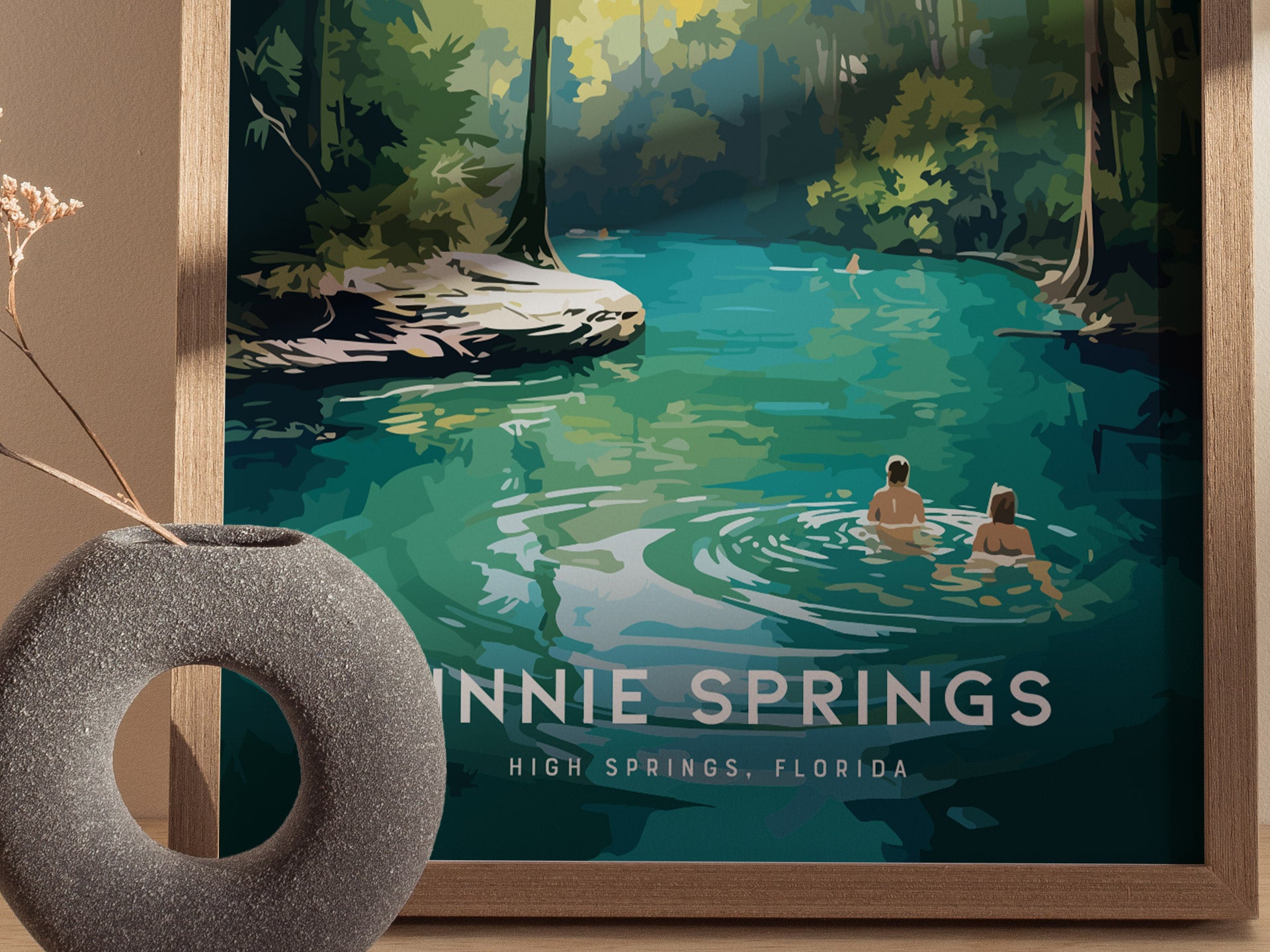A framed poster of Ginnie Springs, Florida, shows two people wading in turquoise water surrounded by lush greenery. A textured stone vase with dried flowers is in the foreground.