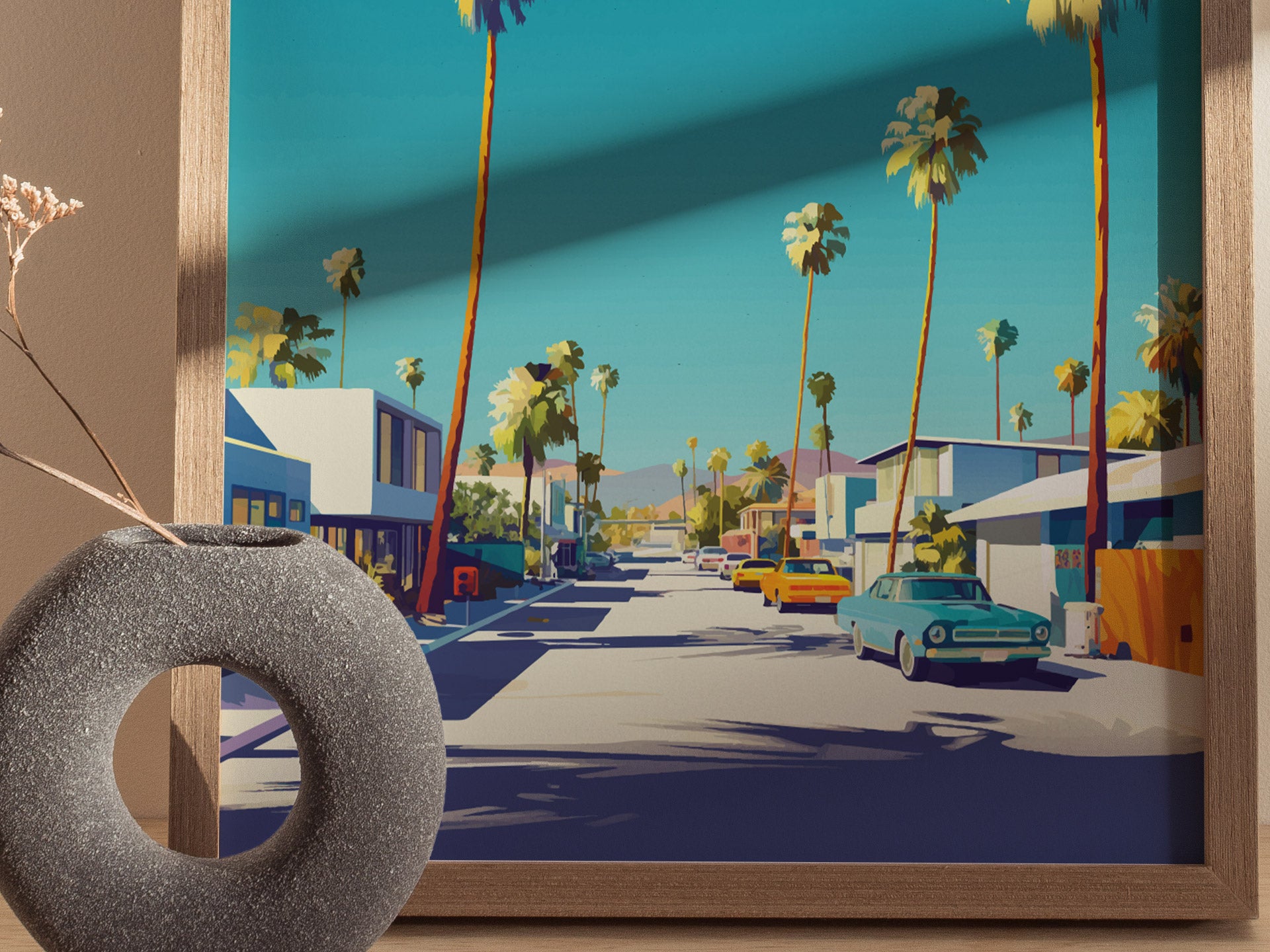 A framed poster depicting a vibrant street scene with palm trees and classic cars under a blue sky, set on a table next to a round, textured vase.