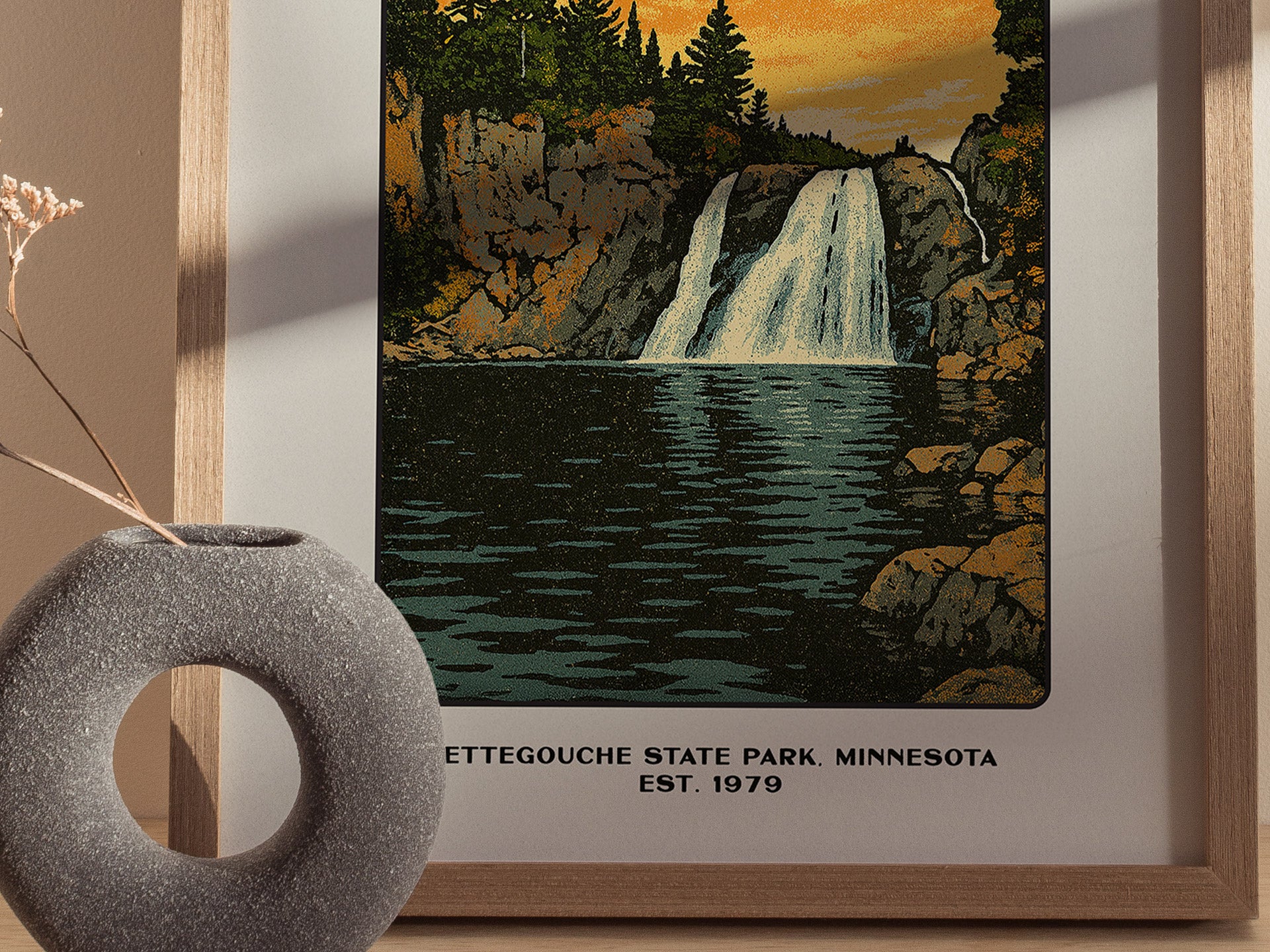 A framed poster of Tettegouche State Park depicts a waterfall cascading between rocky cliffs, with a vase holding dried flowers in the foreground.