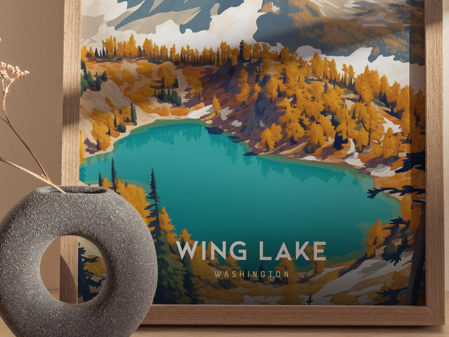 A framed poster of Wing Lake, Washington, featuring vibrant turquoise waters and golden trees, is displayed beside a round stone vase on a wooden surface.