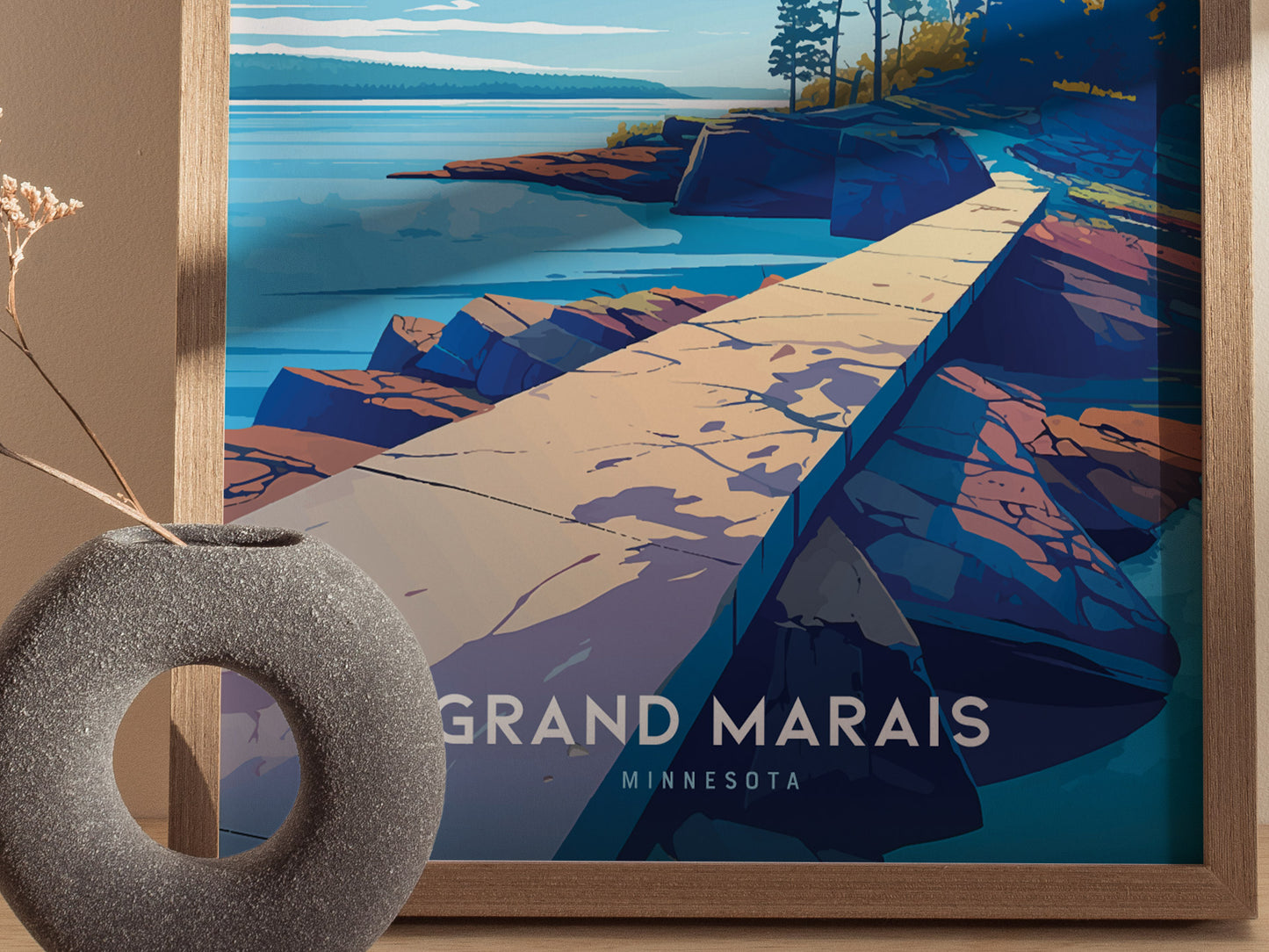 A framed poster depicting a stylized landscape of Artists Point in Grand Marais, Minnesota, features a rocky shoreline and calm waters. A gray circular vase is in the foreground.