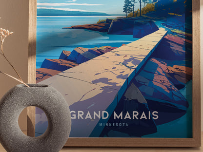 A framed poster depicting a stylized landscape of Artists Point in Grand Marais, Minnesota, features a rocky shoreline and calm waters. A gray circular vase is in the foreground.