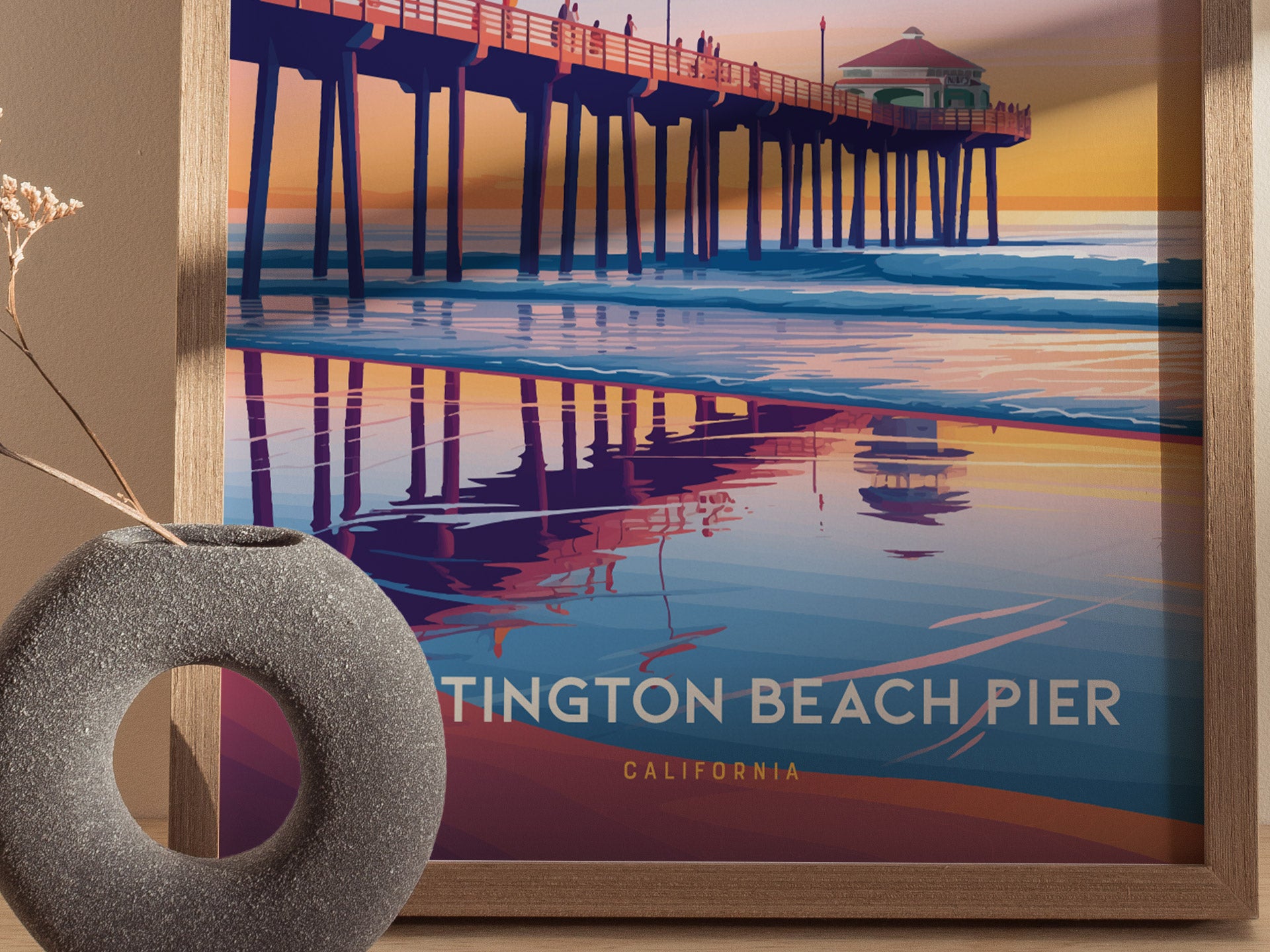 A colorful poster of Huntington Beach Pier at sunset, featuring vivid reflections on the water. The artwork is framed and placed next to a textured, round vase with dry twigs.
