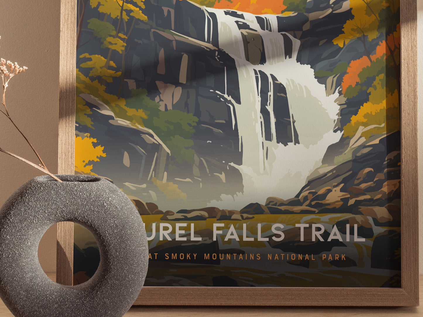 A framed poster depicting a stylized waterfall with autumn foliage in the Great Smoky Mountains, accompanied by a textured gray vase on a wooden surface.