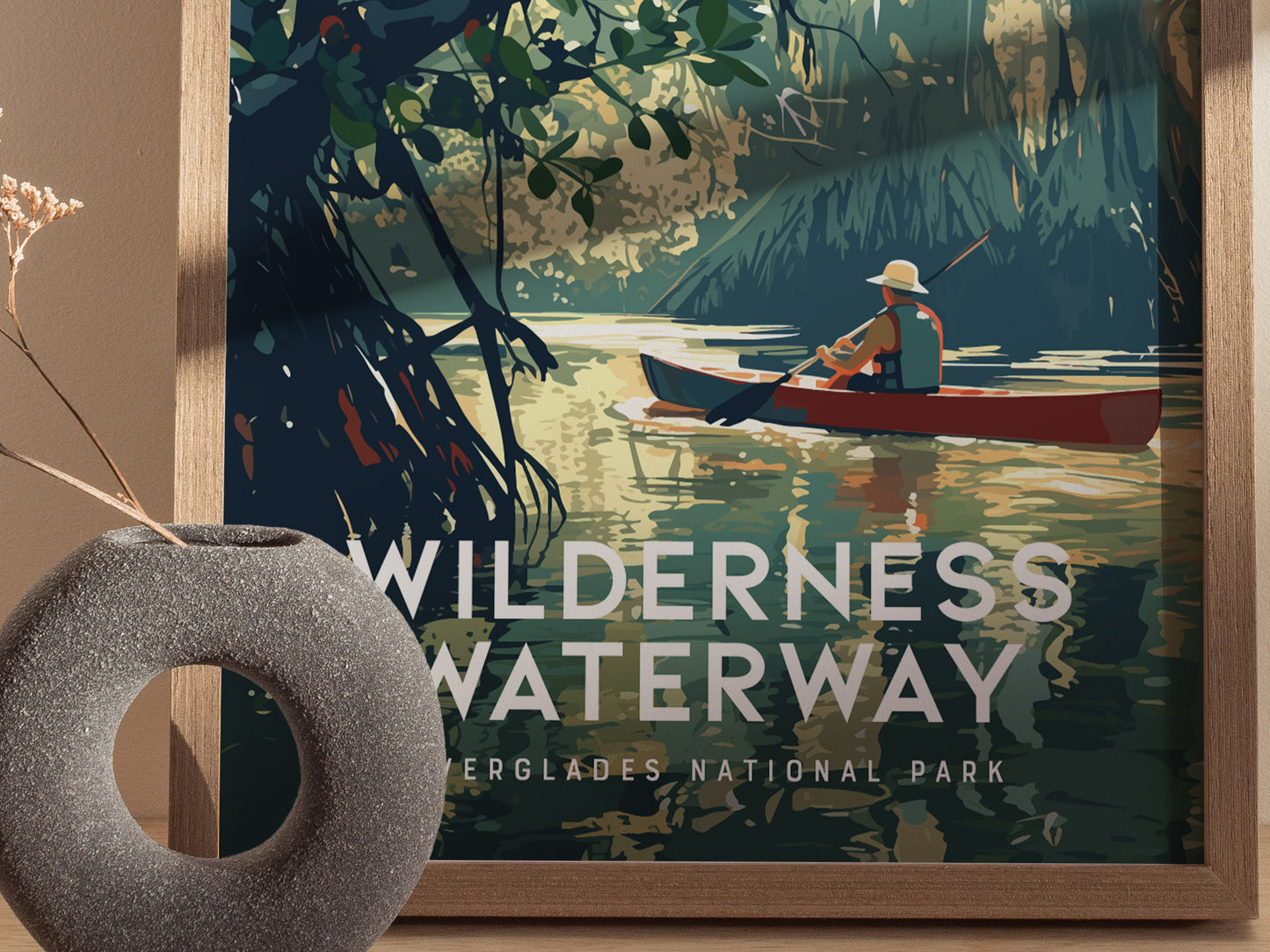 A framed poster titled Wilderness Waterway features a person in a red canoe paddling through lush mangrove forests. A textured gray vase with dried flowers sits in the foreground.
