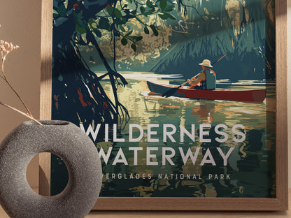 A framed poster titled Wilderness Waterway features a person in a red canoe paddling through lush mangrove forests. A textured gray vase with dried flowers sits in the foreground.