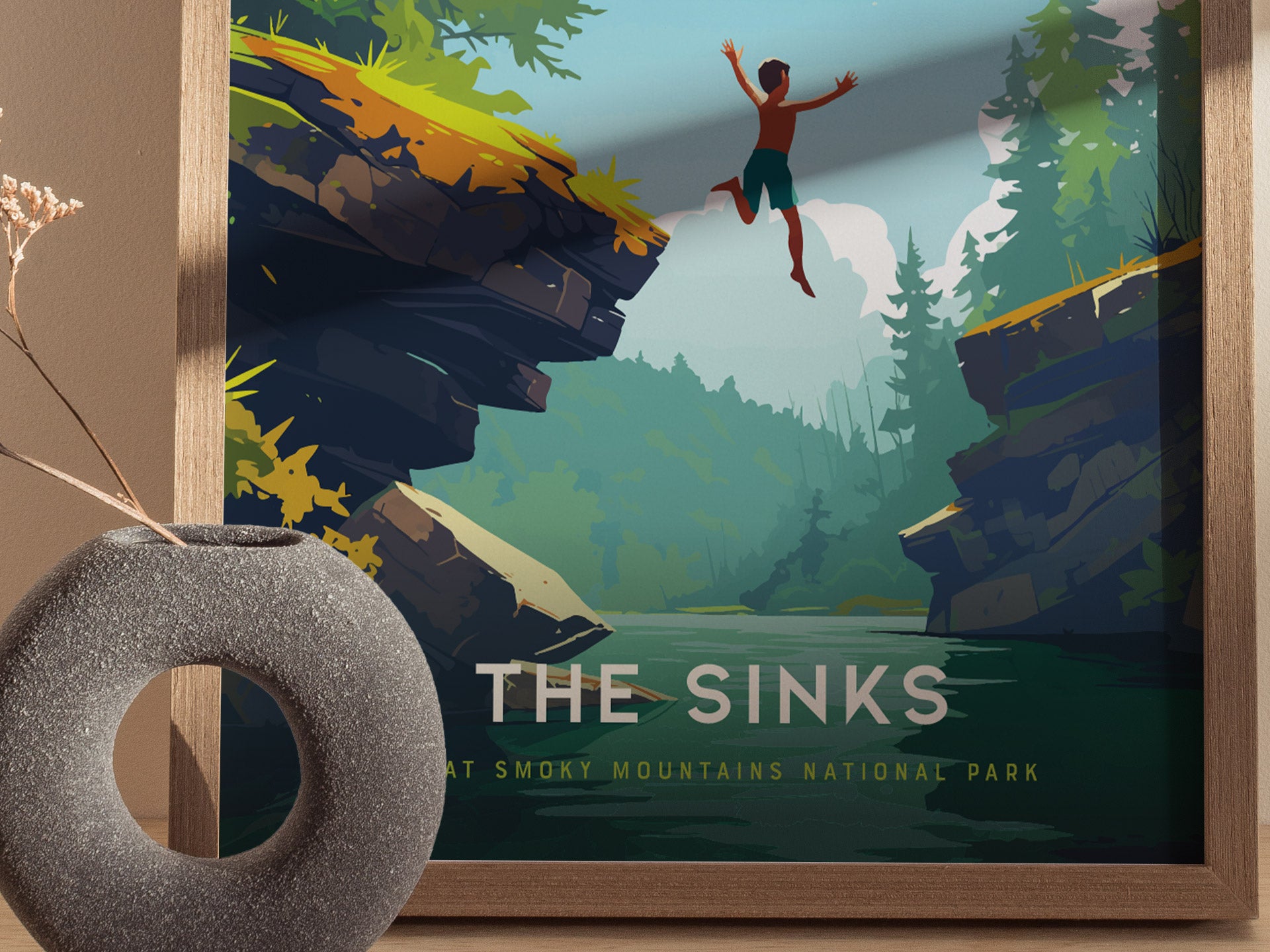 A framed poster titled The Sinks features a vibrant illustration of a person jumping off a rocky ledge into a lush, forested area with a river below. A decorative stone vase with dried flowers is placed in front of the poster.