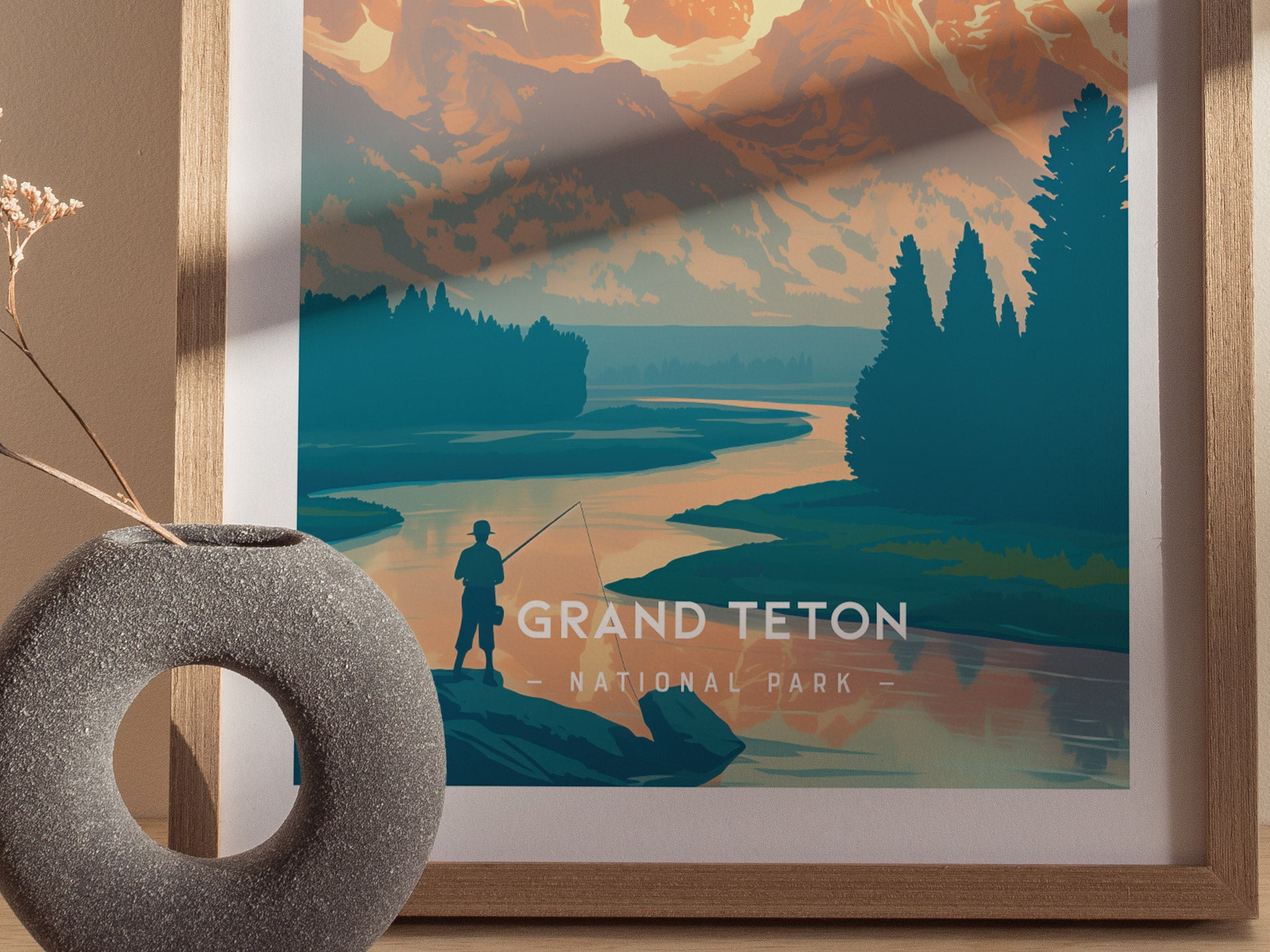 A framed minimalist poster of Grand Teton National Park features a lone fisherman by a tranquil river, with vibrant mountains and trees in the background.