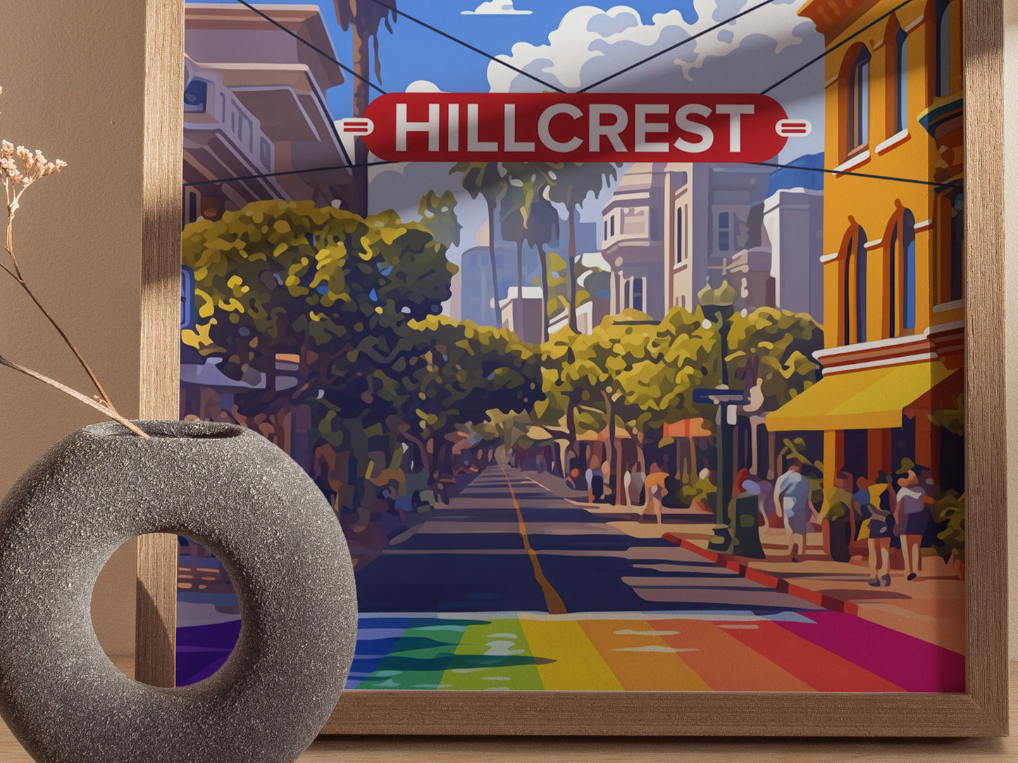 Framed artwork depicts a vibrant street scene in Hillcrest, San Diego, with a prominent red sign and colorful rainbow crosswalk. A gray, donut-shaped vase is in the foreground.
